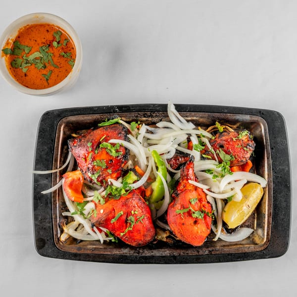 Tandoori chicken with onions, peppers, and lemon on a hot plate, with a side of sauce.