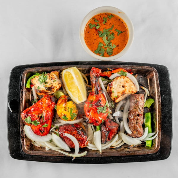 Sizzling platter of mixed grilled meats and vegetables with a side of red sauce.