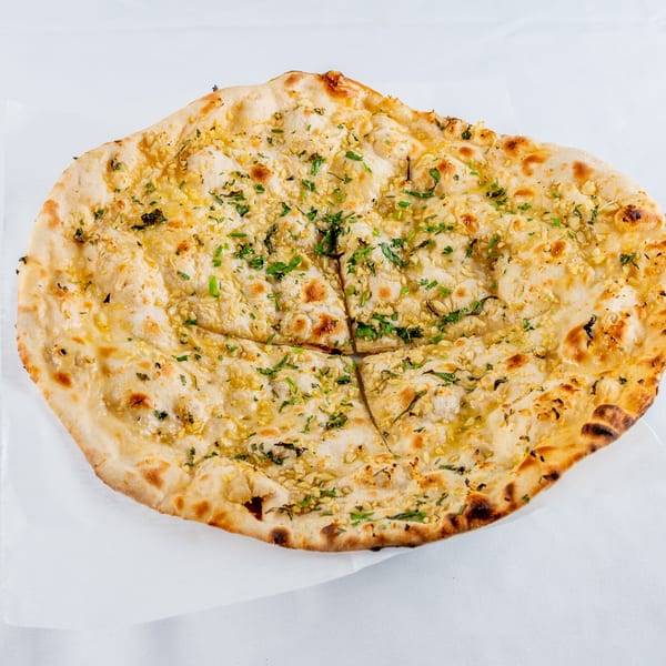 Golden-brown garlic naan bread, topped with minced garlic and fresh green herbs, on a white surface.